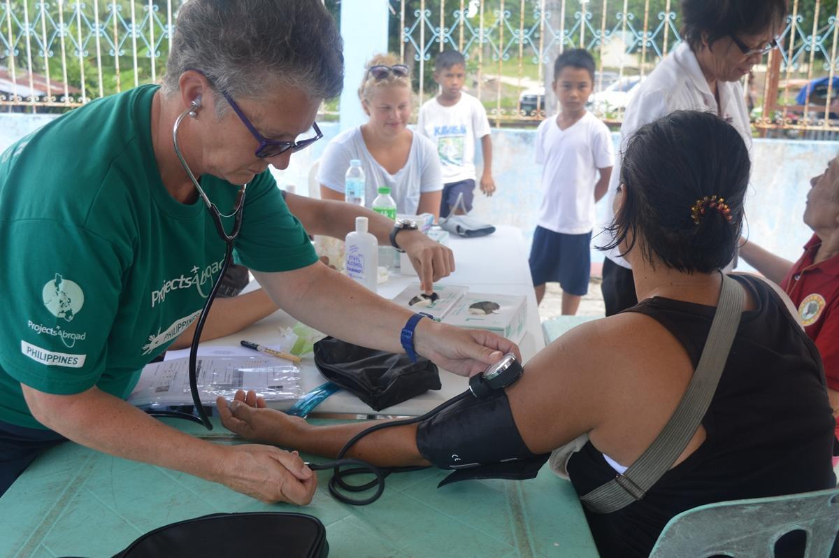 An older adult volunteering in the Philippines checks a woman’s blood pressure during a medical outreach. A woman participating in a volunteer abroad programme for older adults in the Philippines helps a young woman at an outreach